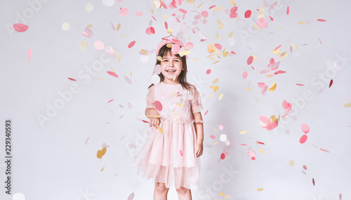 Happy cute little girl wearing pink dress in tulle with princess crown on head isolated on white background playing with confetti. Joyful pretty little girl celebrating her birthday party, having fun