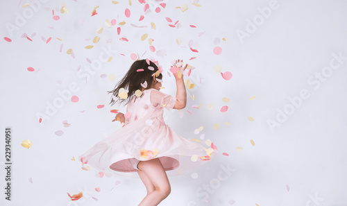 Cheerful cute little girl wearing pink dress in tulle with princess crown dancing on confetti surprise isolated on white studio wall. Playful toddler girl celebrating her birthday party having fun