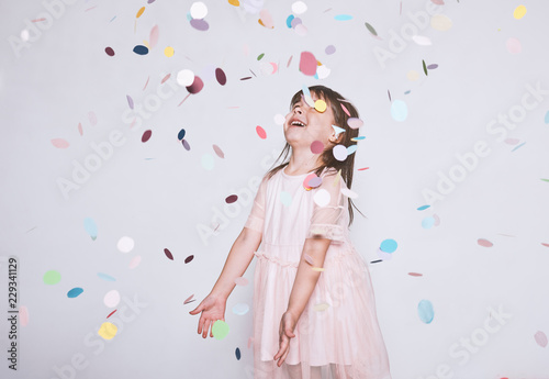 Adorable little girl wearing pink dress in tulle with princess crown on head isolated on white background enjoy confetti surprise. Happy playful girl celebrating her birthday party, having fun