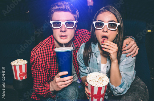 Portrait of two people sitting together in cinema hall and wearing glasses. Girl is amazed and eating popcorn while her partner is drinking cola.