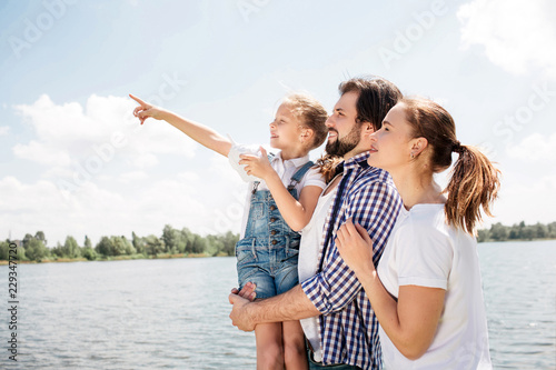 Happy family is standing together and looking up. Guy is holding hi daughter. Girl is pointing up and forward. Woman is standing besides her husband.