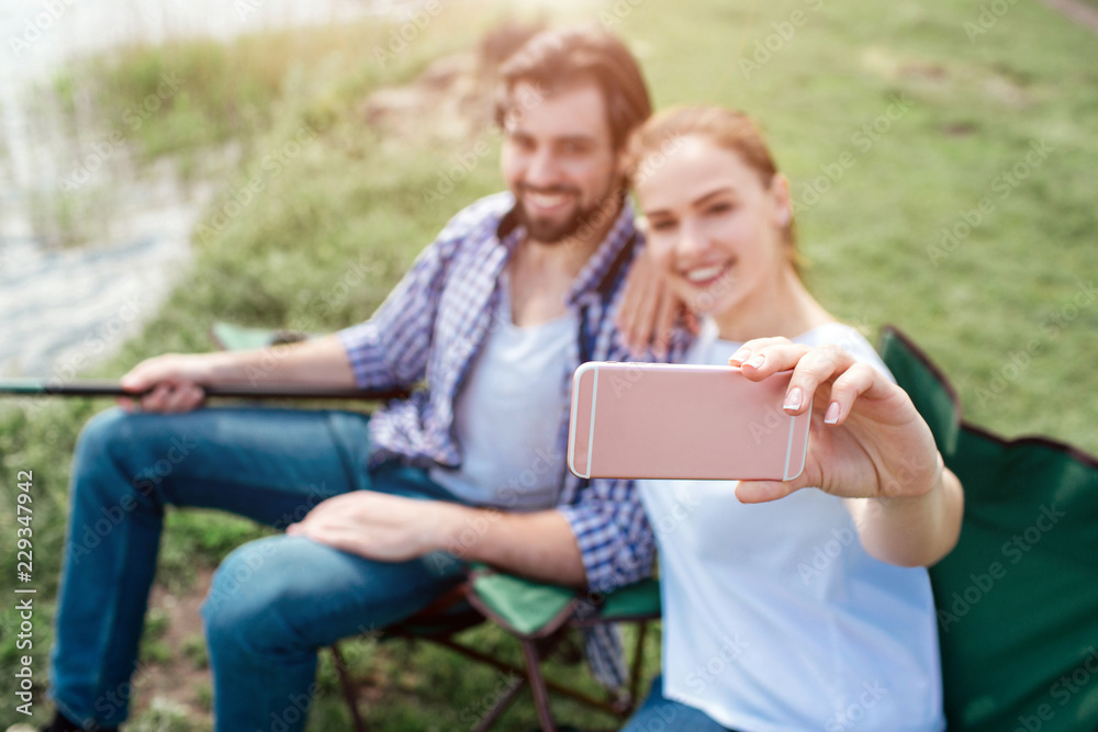 Girl is taking selfie of herself and her husband. They are looking at phone and smiling. Guy is holding the end of fish-rod. People are sitting in folding chairs.