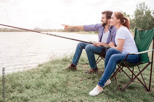 Two people are sitting at river shore. Guy is holding the edge of fish-rod and pointing on water while girl is looking at the same direction as guy is. They are very calm and peaceful.