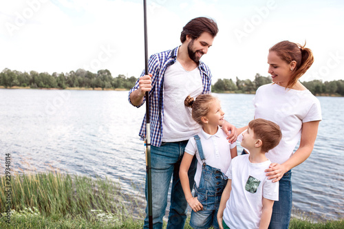 Lovely picture of happy family standing together outside. Guy is holding fish-rod and looking at daughter while she is looking at her mom. Woman is looking at daughter when boy is looking at father.
