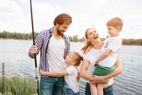 Happy family is standing together. Woman is laughing out loud and holding son in her hands. Small girl is hugging her dad. Guy is holding a fish-rod in right hand.