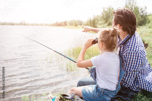 Man is sitting on grass near water with his daughter and pointing forward. Girl is looking there through binoculars. He is holding fish-rod in hands.