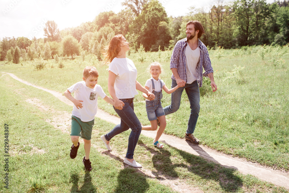 Fototapeta premium A picture of full family walking and jumping together on the road. THey are going through green meadow. They are laughing.