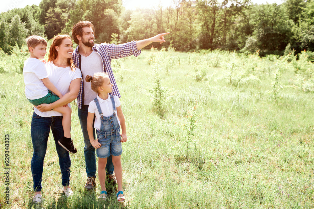 Fototapeta premium Nice picture of family standing together on meadow. Mom is holding her son on hands. Girl is standing besides her parents. Guy is pointing down further.