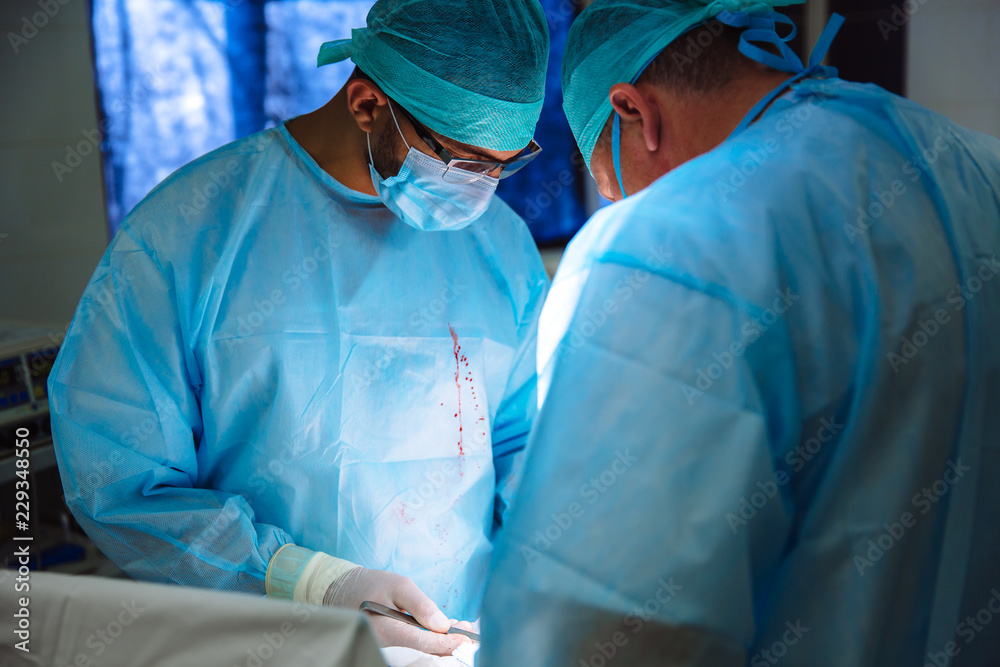 Two doctors in sterile medical clothes perform an operation in a hospital. Professional work in the clinic, surgeons are men with an assistant woman. The concept of health, medicine. Two doctors in sterile medical clothes perform an operation in a hospital. Professional work in the clinic, surgeons are men with an assistant woman. The concept of health, medicine.