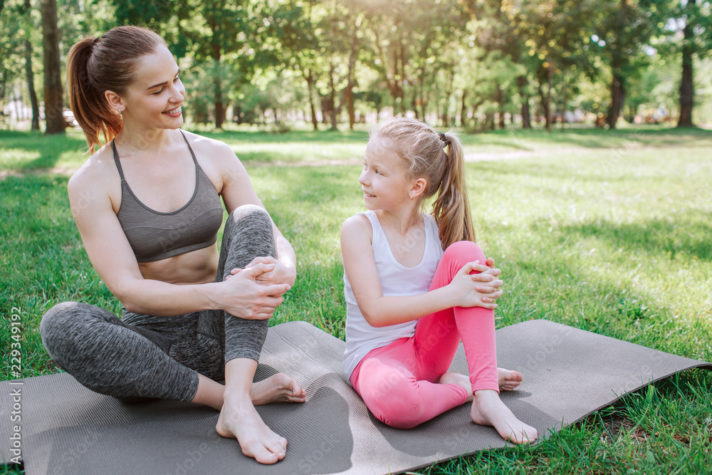 Mom and daughter are sitting on carimate. Their left legs are above right ones. Girls are looking at each other and smiling. They have some rest.