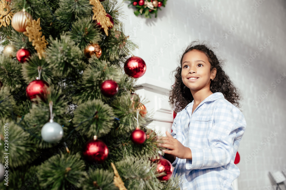 smiling adorable african american child in pajamas decorating christmas tree with baubles and looking away at home
