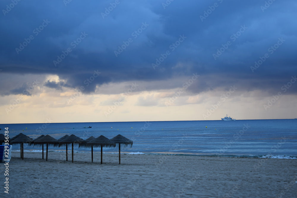 Sandy beach of the Mediterranean coast of Spain with parasols, palm trees and a view of a sailing ship away in cloudy rainy weather Sandy beach of the Mediterranean coast of Spain with parasols, palm trees and a view of a sailing ship away in cloudy rainy weather