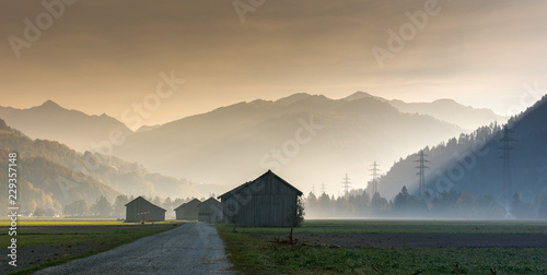 morning mist in a mountain valley with fields and old wooden barns and lattice cross power lines and mountains in silhouette behind