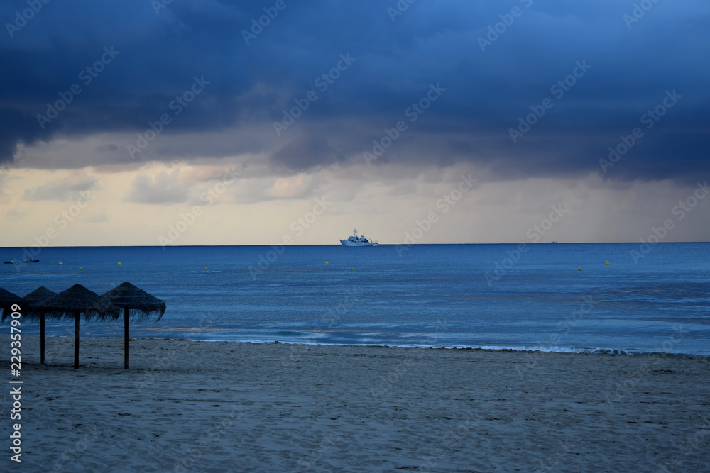 Sandy beach of the Mediterranean coast of Spain with parasols, palm trees and a view of a sailing ship away in cloudy rainy weather Sandy beach of the Mediterranean coast of Spain with parasols, palm trees and a view of a sailing ship away in cloudy rainy weather