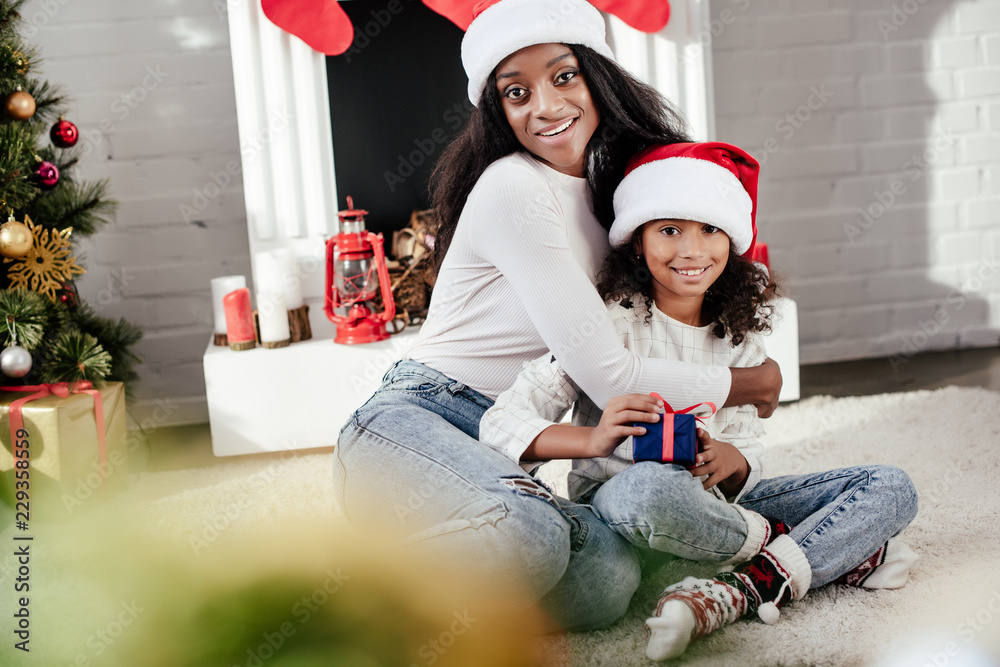 smiling african american woman hugging little daughter with christmas gift in decorated room at home