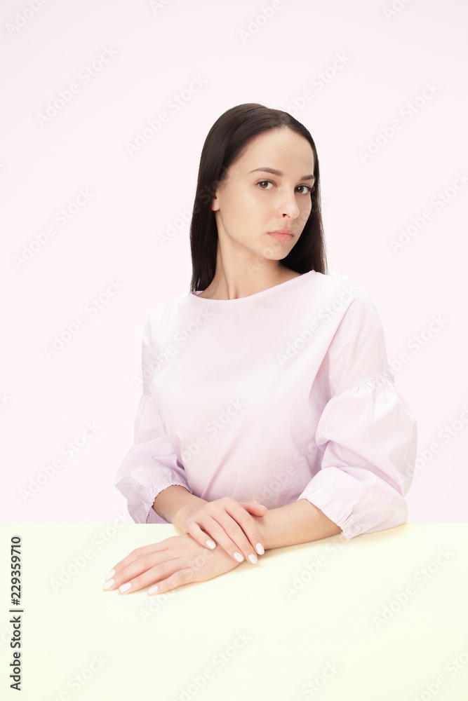 Serious business woman sitting at table on a pink studio background. The portrait in minimalism style Serious business woman sitting at table on a pink studio background. The portrait in minimalism style