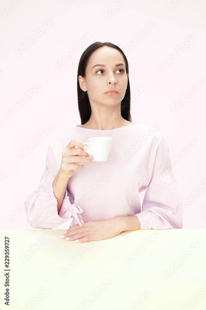 Beautiful lonely woman sitting at pink studio and looking sad holding the cup of coffee in hand. Closeup toned portrait in minimalism style Beautiful lonely woman sitting at pink studio and looking sad holding the cup of coffee in hand. Closeup toned portrait in minimalism style