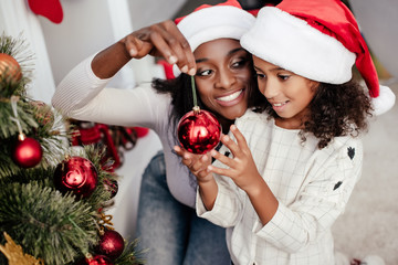 Smiling African American woman in Santa Claus hat helping daughter to decorate Christmas tree at home
