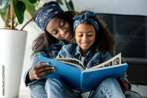 portrait of smiling african american mother and daughter looking at family photo album at home