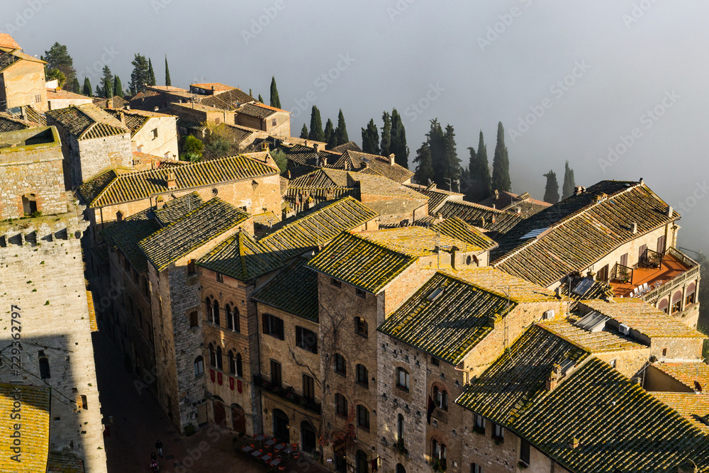 A portion of the little town San Gimignano seen from the top of one of its medieval towers, le limit of the town situated on a hill is marked by a dense white fog 