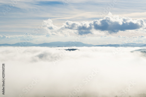 A view from an high tower in the little town of San Gimignano, tuscany italy. A sea made of fog until the horizon with light blue mountains in the background and a little town emerging from the mist  © eugpng