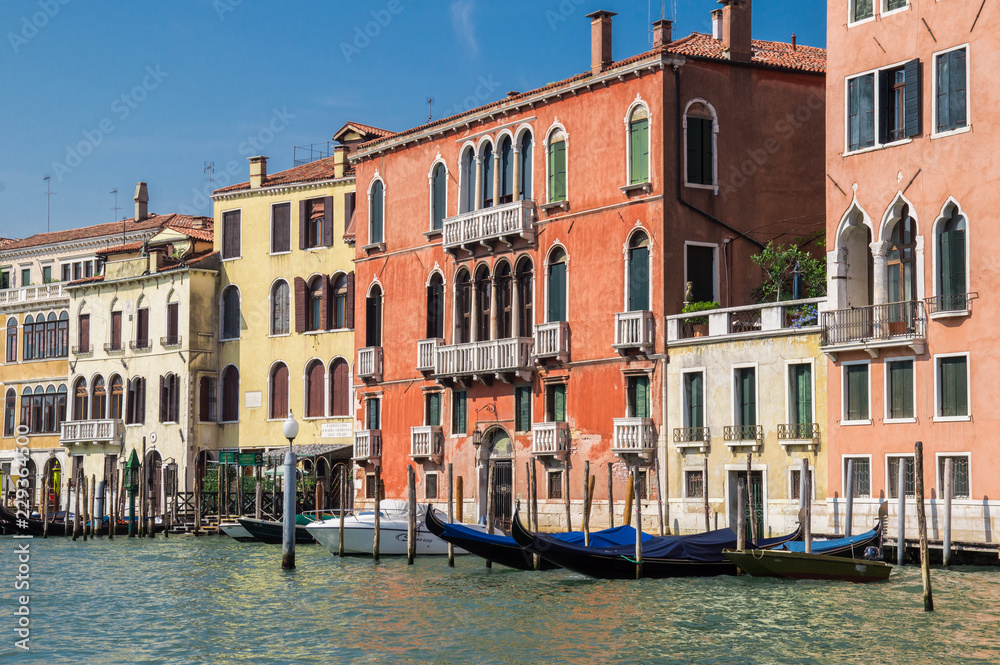 The picturesque embankment of the Grand Canal. Bright Venetian houses on the water, a pier with wooden gondolas. In the background, blue cloudless sky.