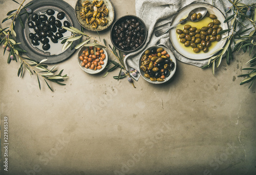 Flat-lay of various kinds of Mediterranean pickled olives in plates and bowls and olive tree branches over grey concrete table background, top view, copy space. Mediterranean meze appetizer
