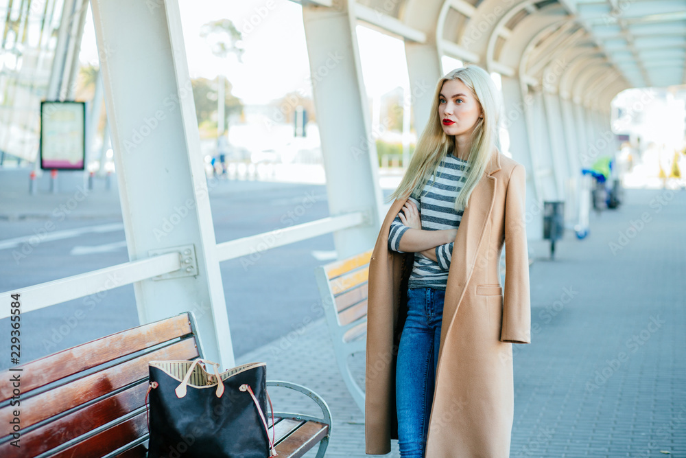 Young charming elegant woman in beige coat waiting for a bus at bus stop. Young charming elegant woman in beige coat waiting for a bus at bus stop.