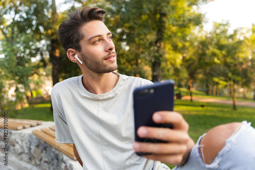 Young guy sitting in park outdoors using mobile phone listening music with earphones.