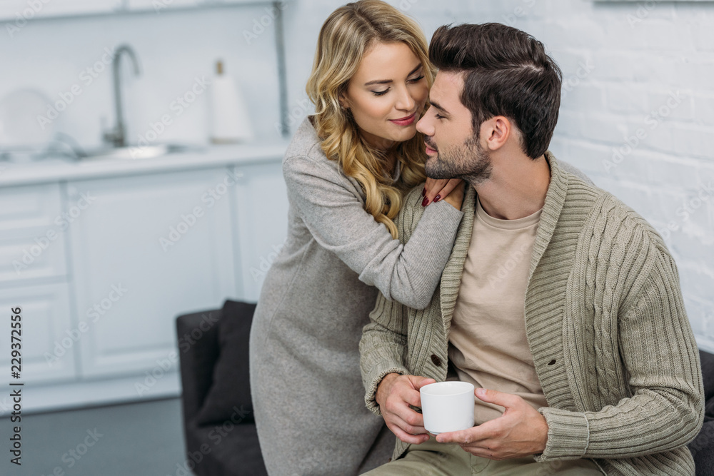 girlfriend hugging boyfriend and he holding cup of tea in kitchen girlfriend hugging boyfriend and he holding cup of tea in kitchen
