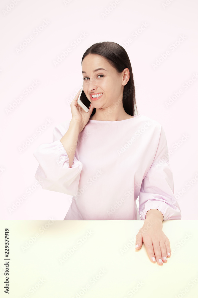 Smiling happy business woman sitting at table isolated on trendy pink studio background with mobile phone. Beautiful, young face. Female half-length portrait. Human emotions, facial expression concept Smiling happy business woman sitting at table isolated on trendy pink studio background with mobile phone. Beautiful, young face. Female half-length portrait. Human emotions, facial expression concept