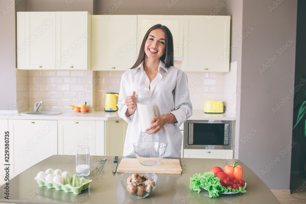 Nice and pretty brunette stands at table with meal and holds glass with milk. She looks on camera and smiles. Girl wears white blouse. Nice and pretty brunette stands at table with meal and holds glass with milk. She looks on camera and smiles. Girl wears white blouse.