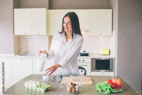 A picture of beutiful girl standing in kitchen and pouring milk in glass cup. She is looking at it and smiling. Girl is concentrated.