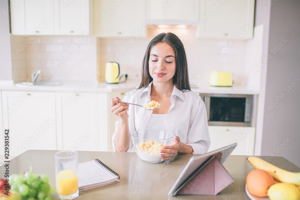 Beautiful young woman sits at table and eats milk with corn flakes. She holds spoon with it. Girl is looking at spoon. There is a notebook with recipes at table and tablet as well.