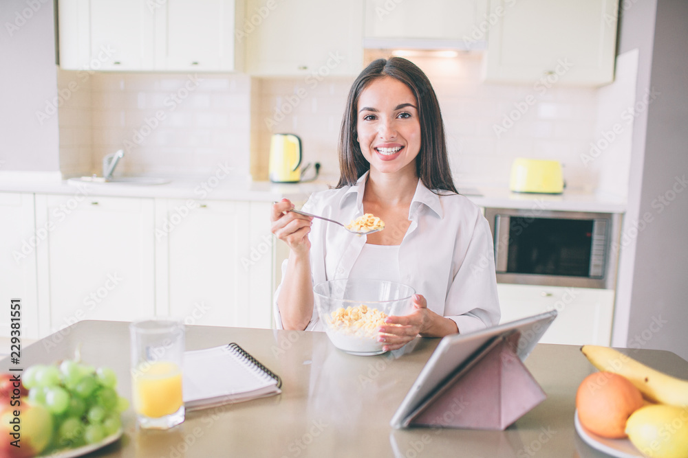 Happy ans positive girl is smiling on camera. She holds a spoon with milk and corn flakes. She is sitting in kitchen.