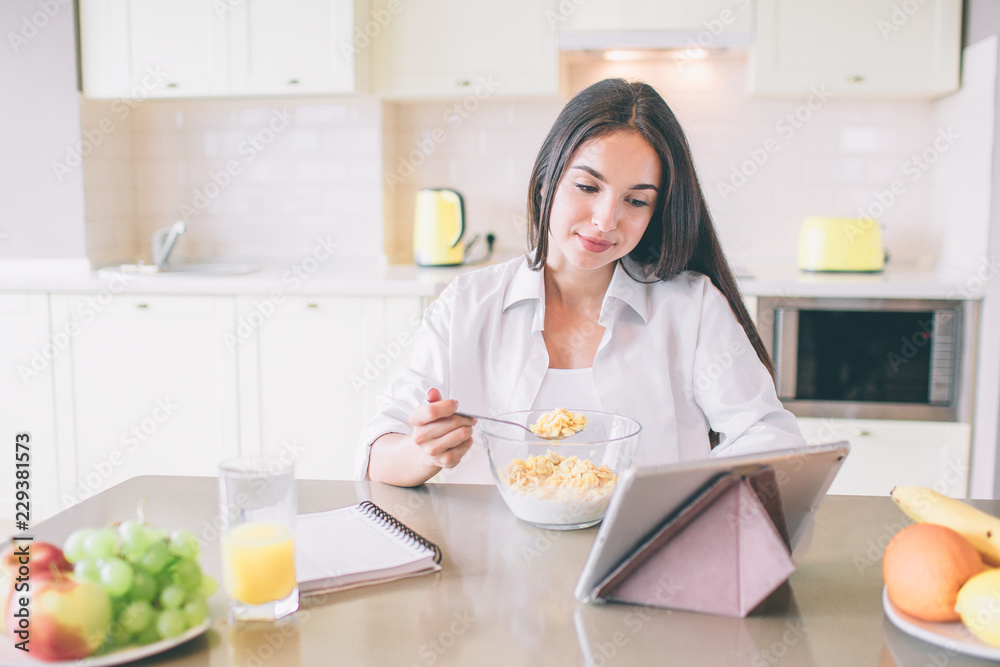 Nice and positive young woman sits at table in kitchen and eats. She watches on tablet and smiling a bit. There is a notebook lying on table with fruit.