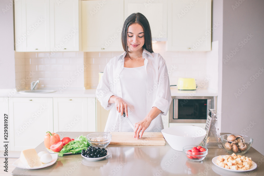 A picture of young woman cooking in kitchen. She stands and cuts vegetable. She is doing that careful.