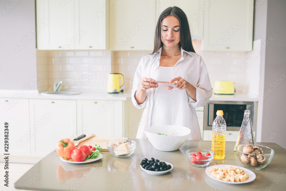 Beautiful girl is sanding in kitchen and looking on the phone she holds in hands. Girl stopped cooking. She has a break.