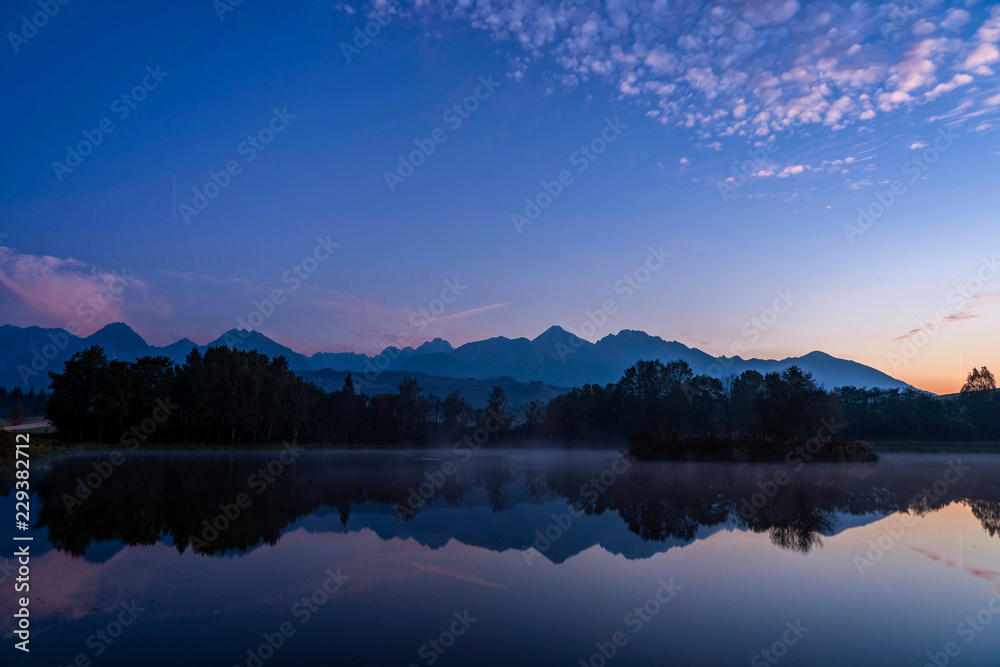 Blue hour shot of peaceful scene of beautiful autumn mountain landscape with lake, colorful trees and high peaks in High Tatras, Slovakia.