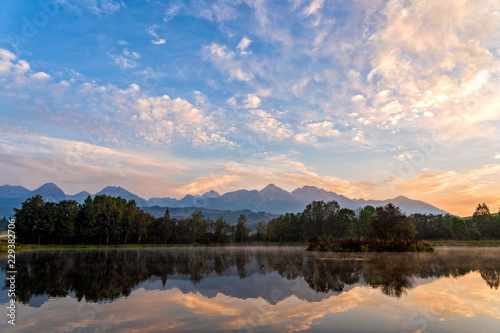 Sunrise shot of peaceful scene of beautiful autumn mountain landscape with lake, colorful trees and high peaks and golden sky in High Tatras, Slovakia.