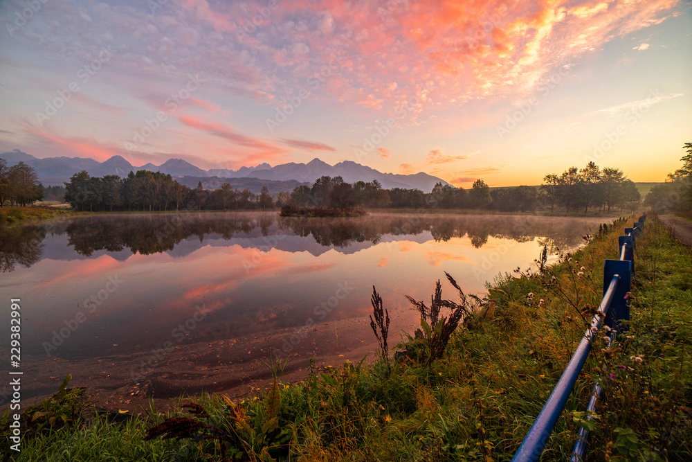 Obraz premium Early morning shot of peaceful scene of beautiful autumn mountain landscape with lake, colorful trees and high peaks and red sky in High Tatras, Slovakia.