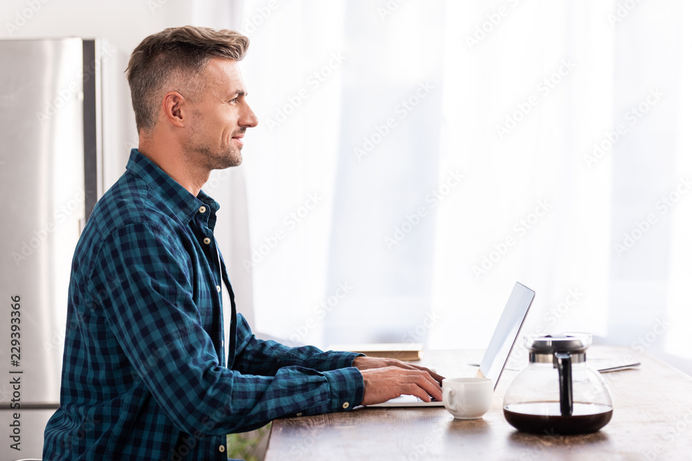 side view of smiling man in checkered shirt using laptop at home