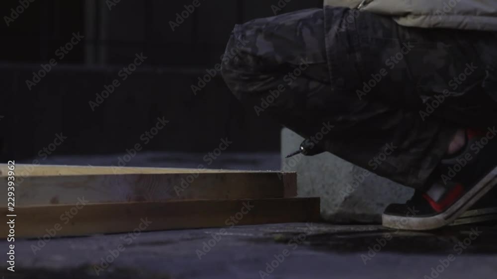 Mature male worker's hands wearing long old grey jacket are using electronic power screw driver to connect two planks placed on ground. Mature male worker's hands wearing long old grey jacket are using electronic power screw driver to connect two planks placed on ground.