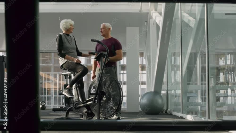 PAN of elderly fitness trainer with grey hair talking to senior woman resting on cycling machine in gym PAN of elderly fitness trainer with grey hair talking to senior woman resting on cycling machine in gym