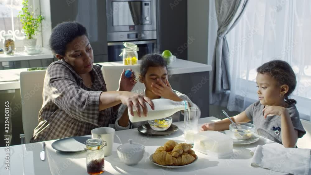 PAN of busy black mother talking on phone and pouring milk into glass for preschool age girl eating breakfast at kitchen table PAN of busy black mother talking on phone and pouring milk into glass for preschool age girl eating breakfast at kitchen table