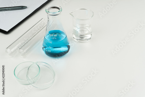 laboratory equipments. A blue liquid flask, clear liquid beaker, test tubes, petri dish and a clipboard with pen on the white table