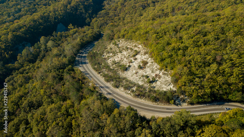Aerial view of curvy rural road in mountains in autumn season. Cars driving below on the road.