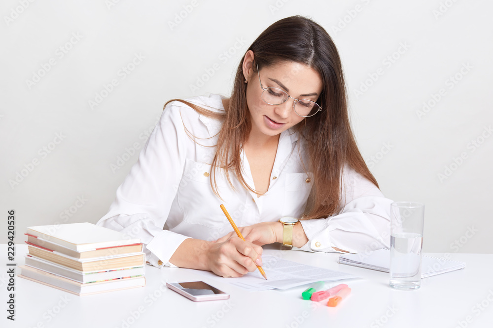 Female entrepreneur writes organisation plan, sits at desktop, uses books and pencil, drinks fresh water from glass, concentrared on work, wears round optical glasses, isolated over white background Female entrepreneur writes organisation plan, sits at desktop, uses books and pencil, drinks fresh water from glass, concentrared on work, wears round optical glasses, isolated over white background