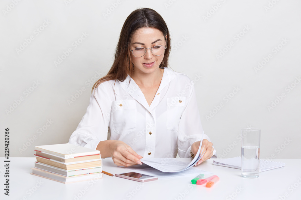 Photo of female enterprise holds paper documents, poses in coworking space, drinks water from glass, wears spectacles for good vision. Busy student writes diploma paper, prepares for final exam Photo of female enterprise holds paper documents, poses in coworking space, drinks water from glass, wears spectacles for good vision. Busy student writes diploma paper, prepares for final exam