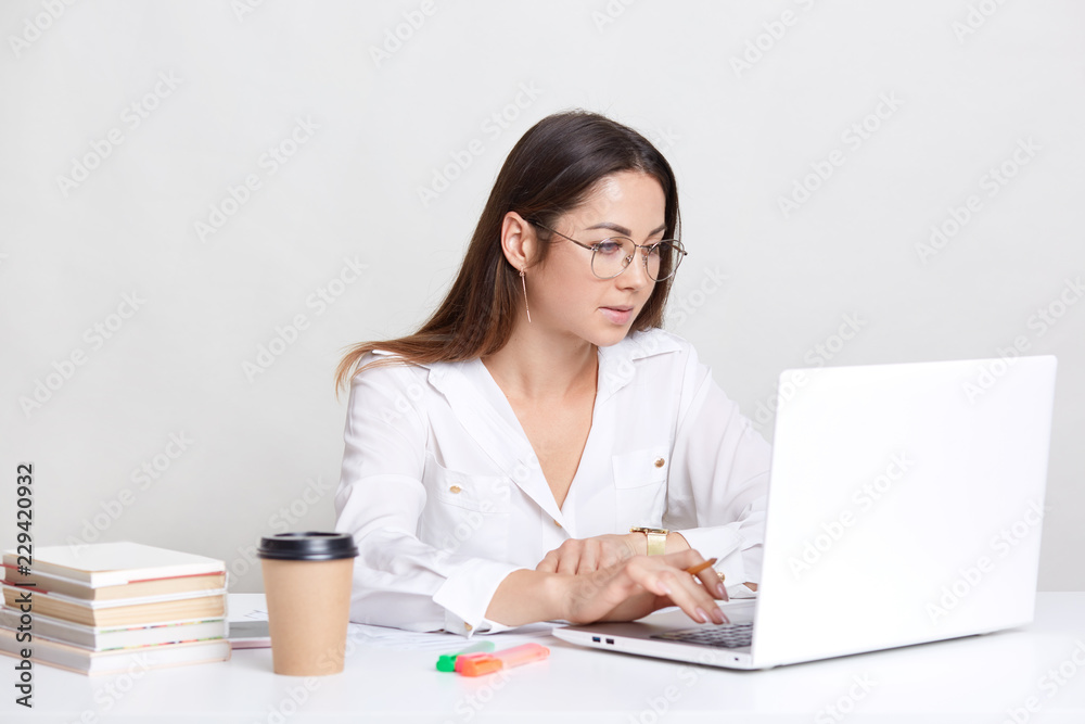Young female administrative works on laptop computer, searches information in internet, dressed in white drinks hot beverage, uses books for writing diploma paper, isolated over white background Young female administrative works on laptop computer, searches information in internet, dressed in white drinks hot beverage, uses books for writing diploma paper, isolated over white background